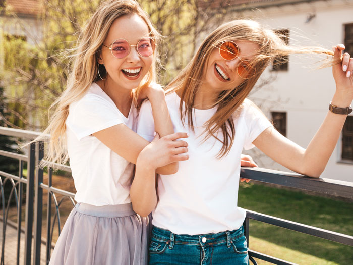Two young women wearing sunglasses and white shirts, smiling and posing together outdoors on a balcony. Two young women wearing sunglasses and white shirts, smiling and posing together outdoors on a balcony.