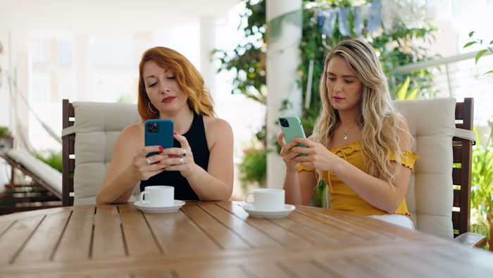 Two women sitting apart at a table, both focused on their phones, reflecting tension between bestie and wannabe influencer. Two women sitting apart at a table, both focused on their phones, reflecting tension between bestie and wannabe influencer.