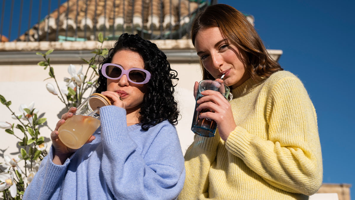 Two women outdoors sipping drinks, one wearing purple sunglasses and a blue sweater, the other in a yellow sweater, casual setting.
