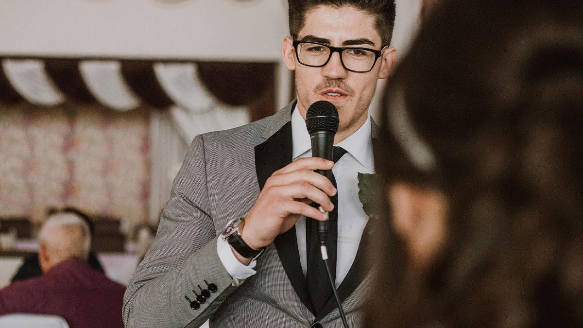 Man giving best man speech at wedding, wearing glasses and suit, holding microphone while guests listen nearby.