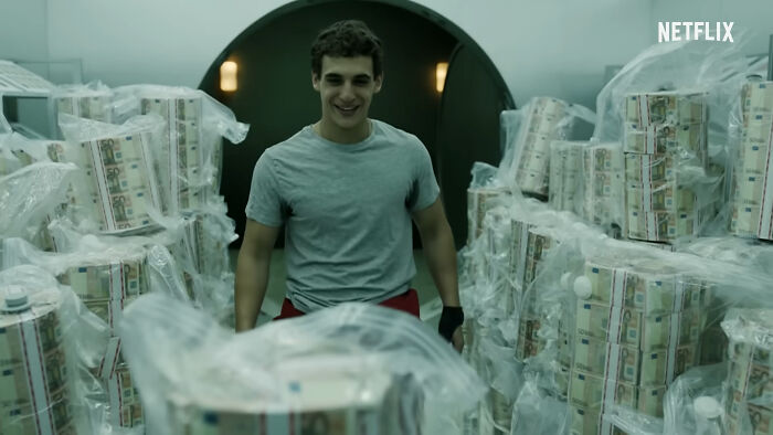Young man smiling amid stacks of wrapped money bundles in a scene from one of the best Netflix shows.