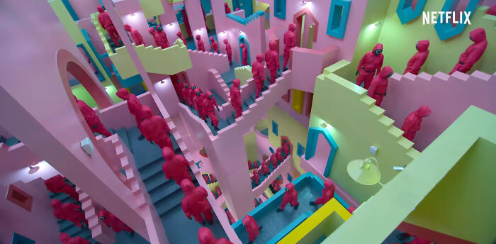 Crowd of red-suited characters navigating colorful, confusing staircases in a popular Netflix show scene.
