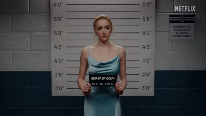 Woman in a blue dress holding a mugshot sign in front of a height chart, from a popular Netflix show series.