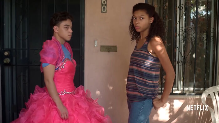 Two young people standing outside a house, one wearing a bright pink dress, from popular Netflix shows.