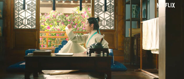 Scene from a popular Netflix show featuring a person in traditional clothing sitting by a low table in a decorated room.