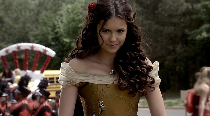 Young woman with long curly hair in a vintage dress at an outdoor event with people and a band in the background.