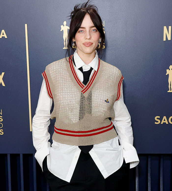Billie Eilish posing at an event, wearing a sleeveless sweater vest over a white shirt and black tie. Billie Eilish posing at an event, wearing a sleeveless sweater vest over a white shirt and black tie.