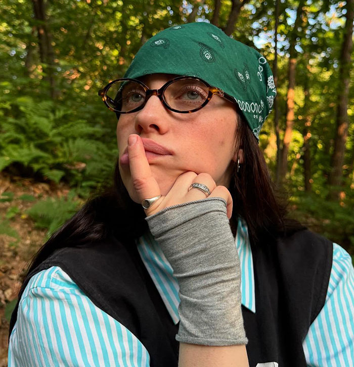 Billie Eilish wearing glasses and green bandana, posing thoughtfully outdoors with trees in the background Billie Eilish wearing glasses and green bandana, posing thoughtfully outdoors with trees in the background