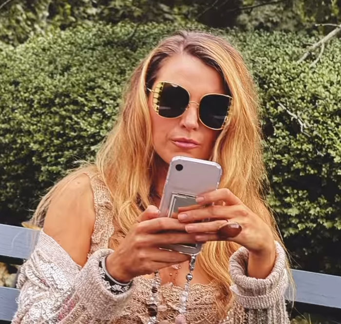Blake Lively wearing large sunglasses outdoors, focused on her phone in a casual setting with greenery behind her. Blake Lively wearing large sunglasses outdoors, focused on her phone in a casual setting with greenery behind her.