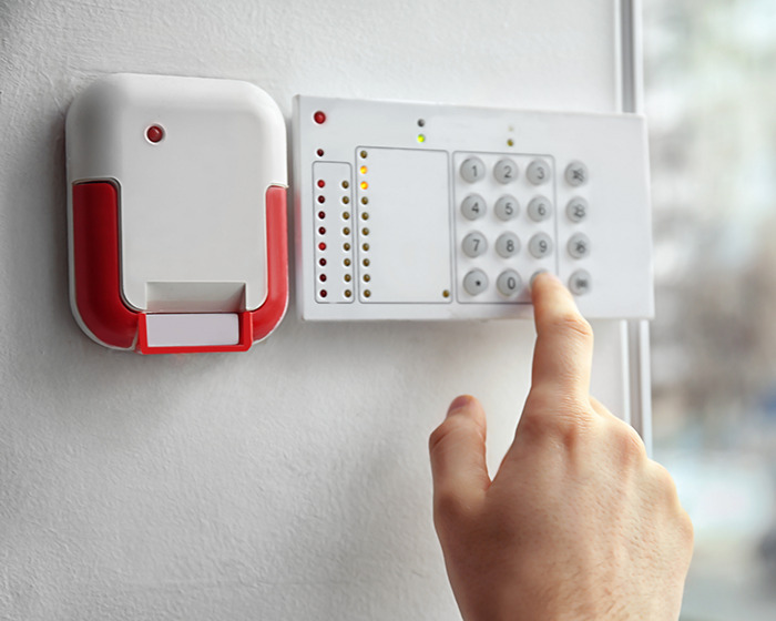 Hand pressing numbers on a security alarm keypad beside a red and white wall-mounted fire alarm system. Hand pressing numbers on a security alarm keypad beside a red and white wall-mounted fire alarm system.