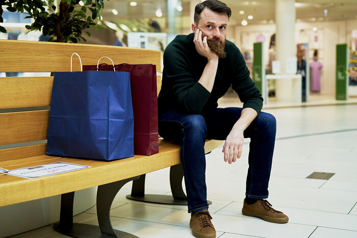 Man sitting on bench in shopping mall looking bored with shopping bags beside him, illustrating public behavior moments.