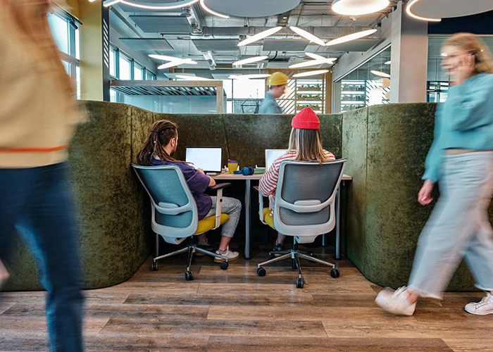 Two employees working on laptops in an office pod with blurred coworkers walking by, illustrating data fudging consequences. Two employees working on laptops in an office pod with blurred coworkers walking by, illustrating data fudging consequences.