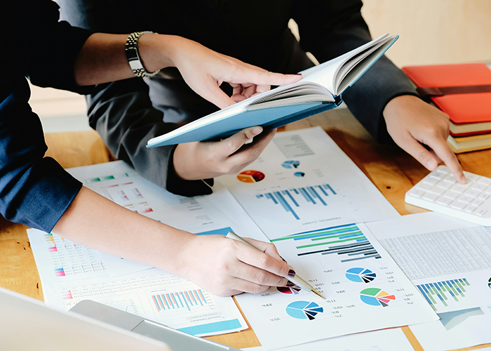 Two colleagues analyzing charts and data reports at a desk, discussing data fudge to improve department results. Two colleagues analyzing charts and data reports at a desk, discussing data fudge to improve department results.