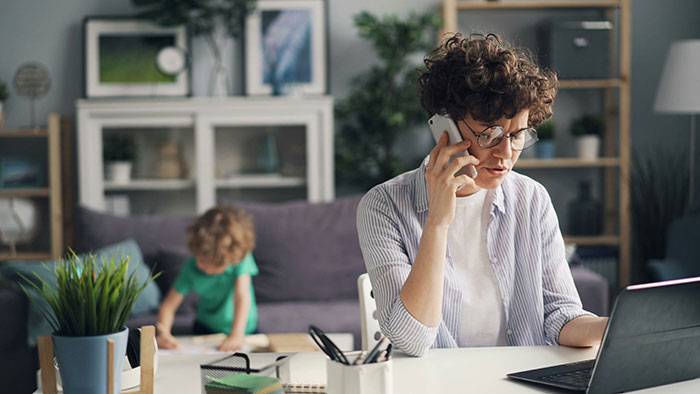 Woman talking on phone while working on laptop at home, with child in background near couch, holding phone to charge. Woman talking on phone while working on laptop at home, with child in background near couch, holding phone to charge.