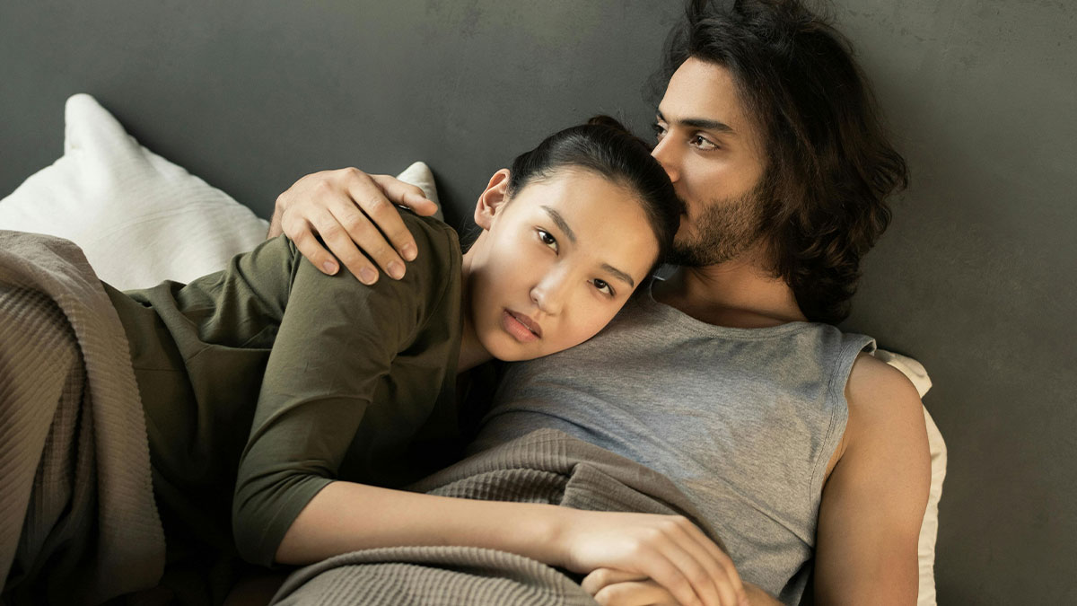 Couple lying together in bed in a bathroom setting, boyfriend holding ex-gf while phone charges nearby.