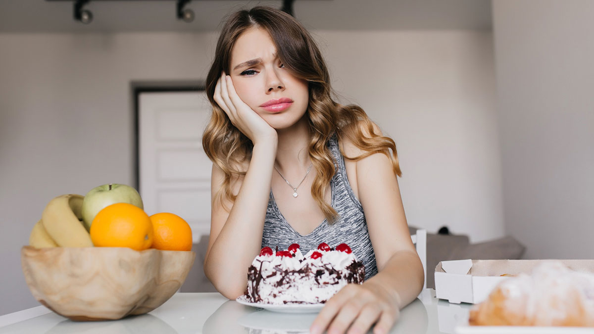Young woman looks upset while sitting at a table with a cake, illustrating guy nagging his starving girlfriend to eat more.