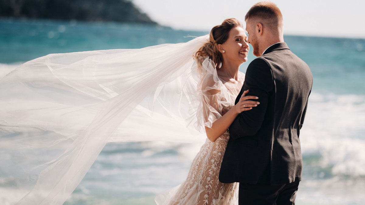 Bride and groom embrace on a windy beach wedding, bride wearing a flowing lace gown with long veil waving in heat.