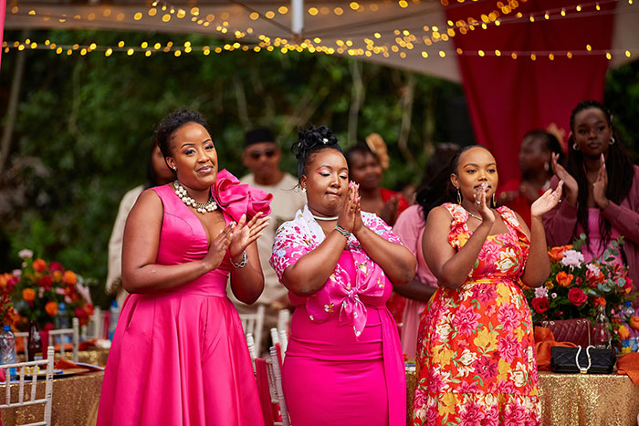 Three women at a wedding celebration, dressed in bright dresses, clapping and smiling with guests in the background. Three women at a wedding celebration, dressed in bright dresses, clapping and smiling with guests in the background.