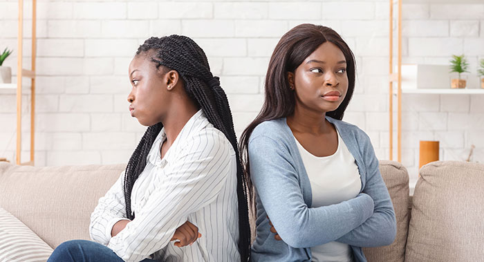 Two women sitting back to back on a couch with arms crossed, showing tension and conflict after money was caught on camera. Two women sitting back to back on a couch with arms crossed, showing tension and conflict after money was caught on camera.