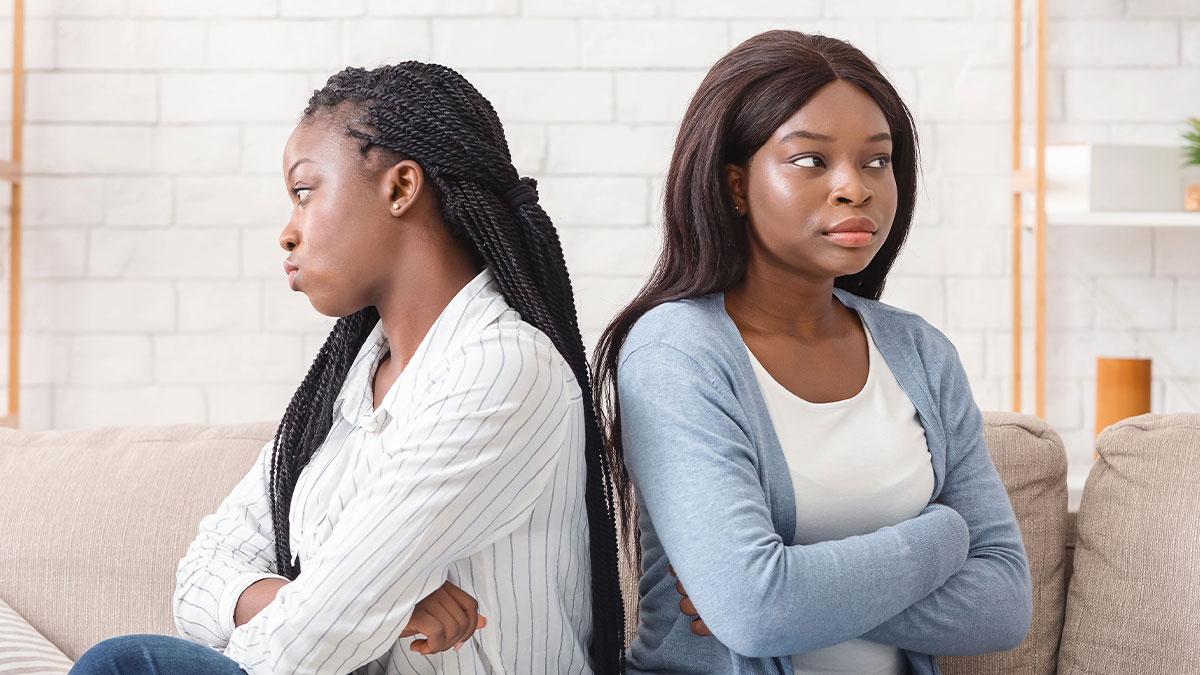 Two women sitting back-to-back on a couch, upset and avoiding eye contact, depicting bride and sister conflict.