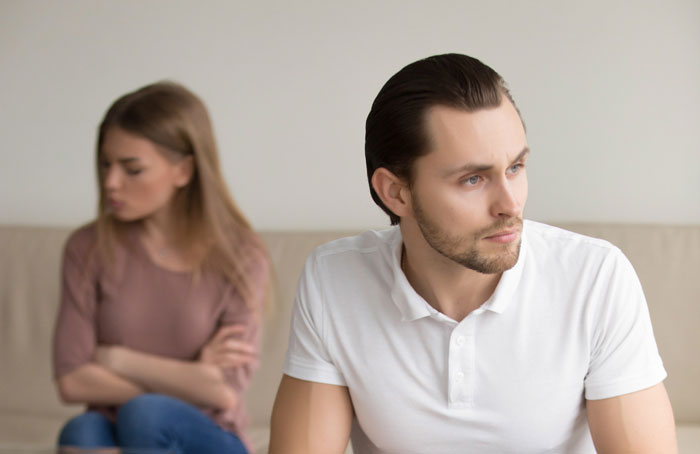 Upset man in white shirt looking away as woman sits behind him with crossed arms, highlighting secret kid and family conflict.