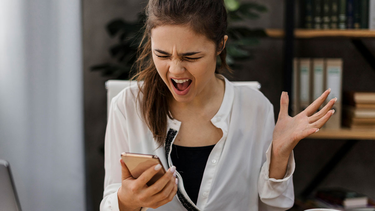 Young woman shocked and frustrated by brutal rejection message seen on her smartphone at home office.