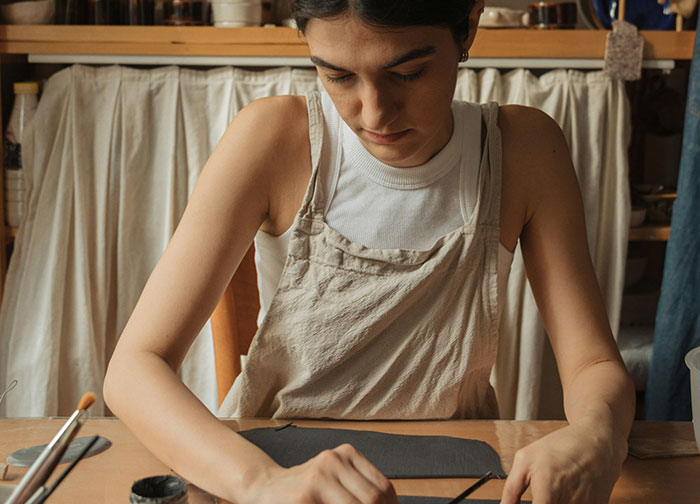 Young woman focused on painting pottery at a wooden table, highlighting men's brutal honesty and personal expression.