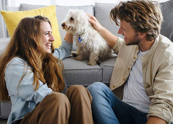 A man and woman sitting on the floor smiling and petting a small white dog, reflecting men brutally honest thoughts.