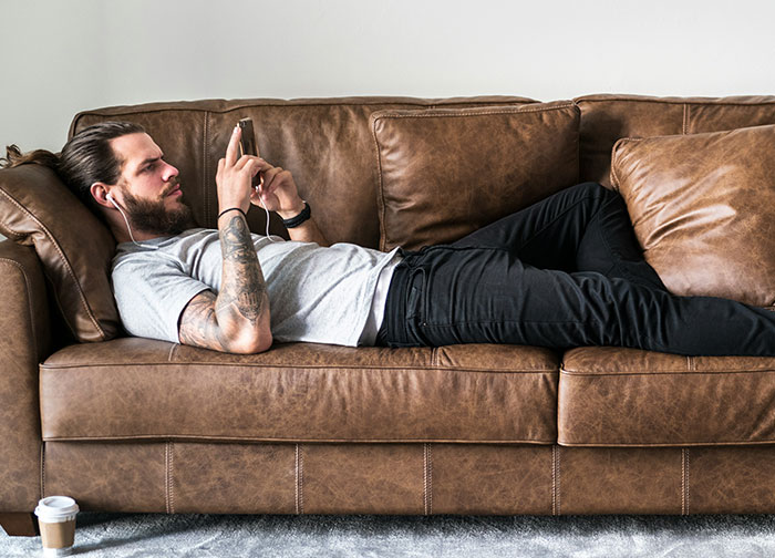 Man lying on brown leather couch using smartphone and wearing earphones, reflecting on things men never tell women.