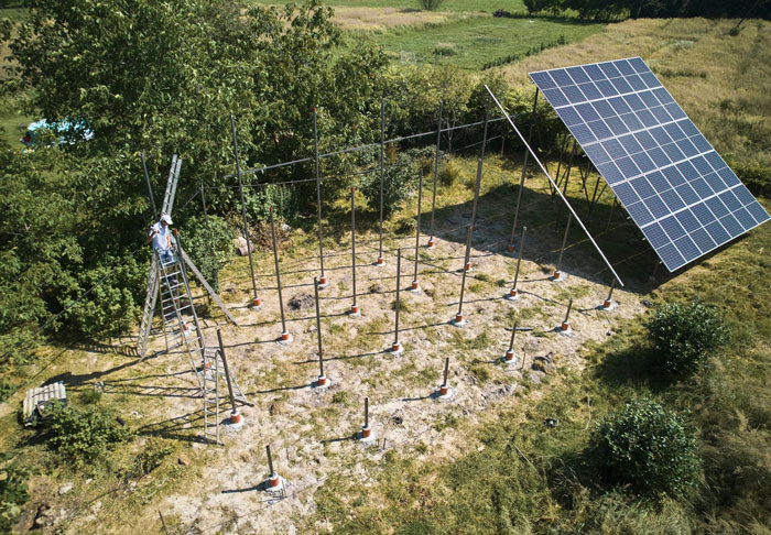 Workers installing solar farm supports in a grassy field next to a large solar panel and surrounding trees. Workers installing solar farm supports in a grassy field next to a large solar panel and surrounding trees.