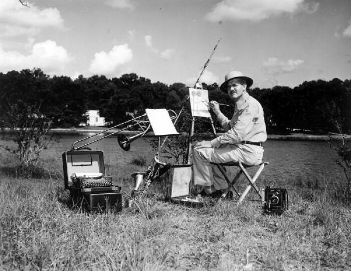 Man painting outdoors by a river with typewriter and trumpet nearby, capturing everyday life in 1950s Florida.