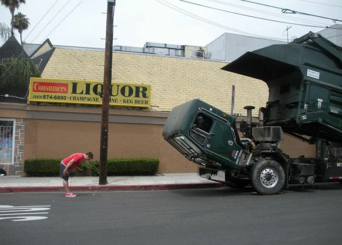Man bending forward next to a large truck with its cab awkwardly tilted upward near a convenience store.