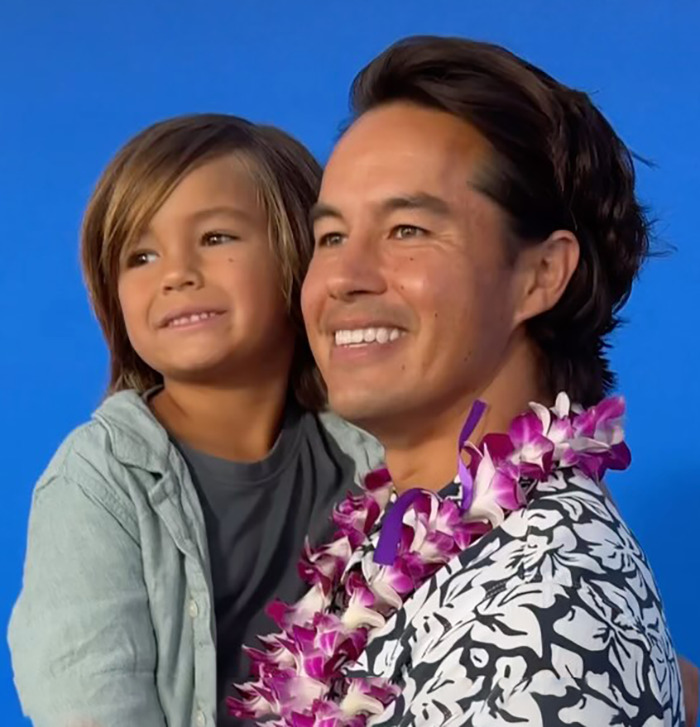 Smiling dad wearing a floral lei holding his young son against a blue background, highlighting father and son relationship fears. Smiling dad wearing a floral lei holding his young son against a blue background, highlighting father and son relationship fears.