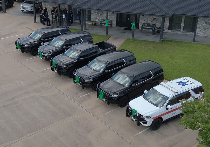 Line of black hearses and an emergency vehicle parked outside a building with people gathered nearby for a memorial event. Line of black hearses and an emergency vehicle parked outside a building with people gathered nearby for a memorial event.