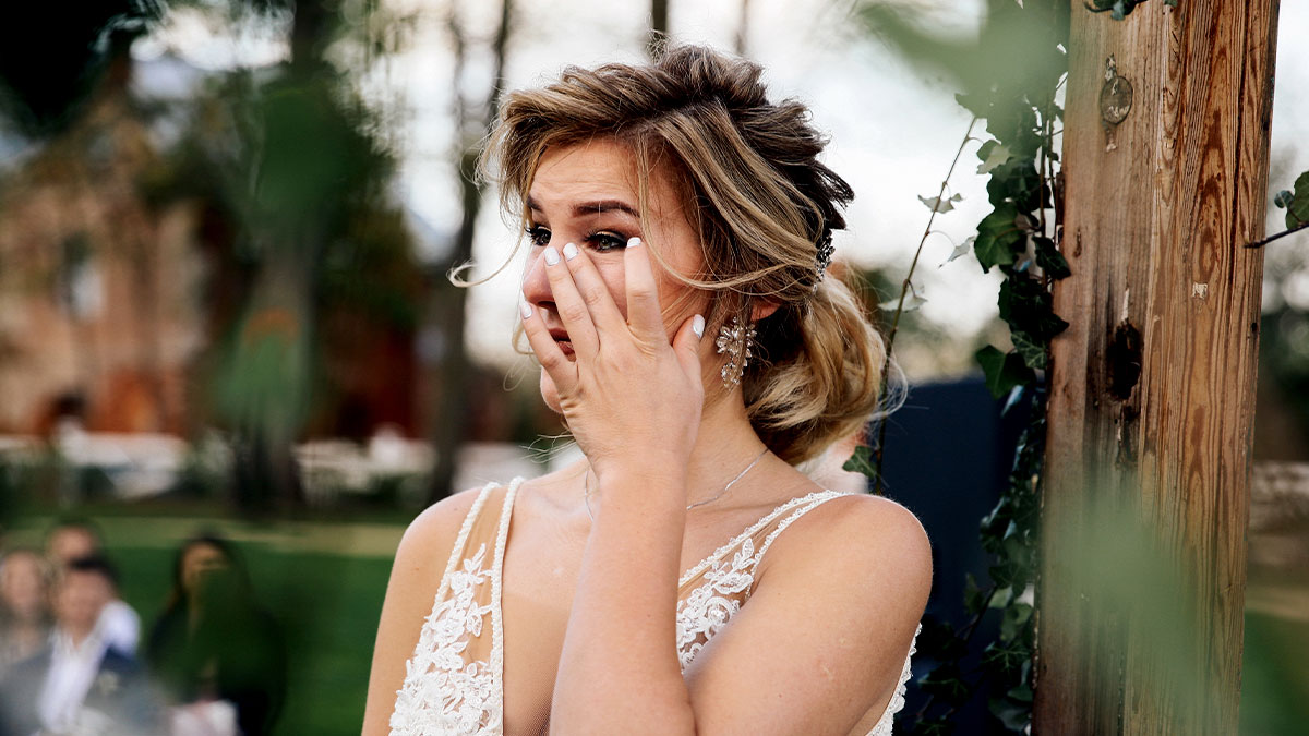 Bride emotional outdoors, wiping tears, wearing wedding dress, upset after caterers got the wedding date wrong before ceremony.