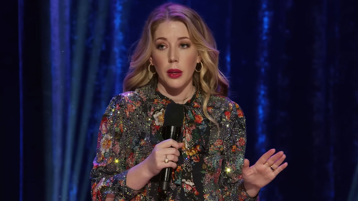 Female comedian performing stand-up on stage holding a microphone with a floral patterned outfit and blue curtain background