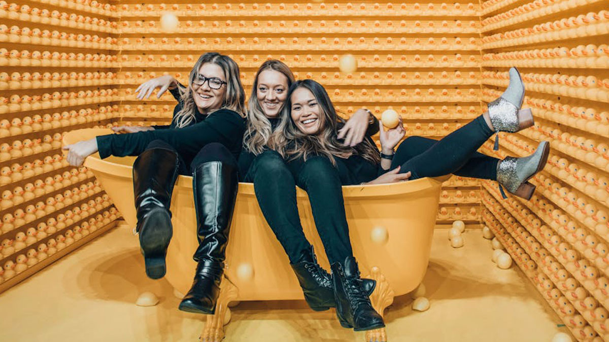 Three women sitting in a yellow bathtub surrounded by rubber ducks in a playful stay hydrated themed room.