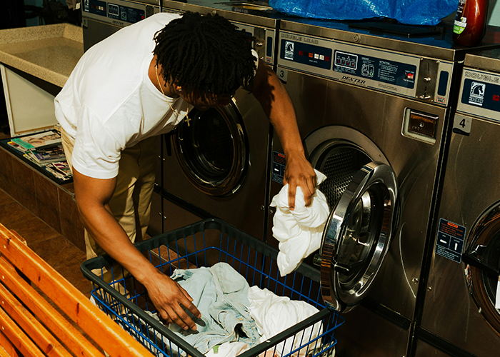 College student loading laundry in a washing machine at a laundromat, showing responsibility and cleaning efforts. College student loading laundry in a washing machine at a laundromat, showing responsibility and cleaning efforts.