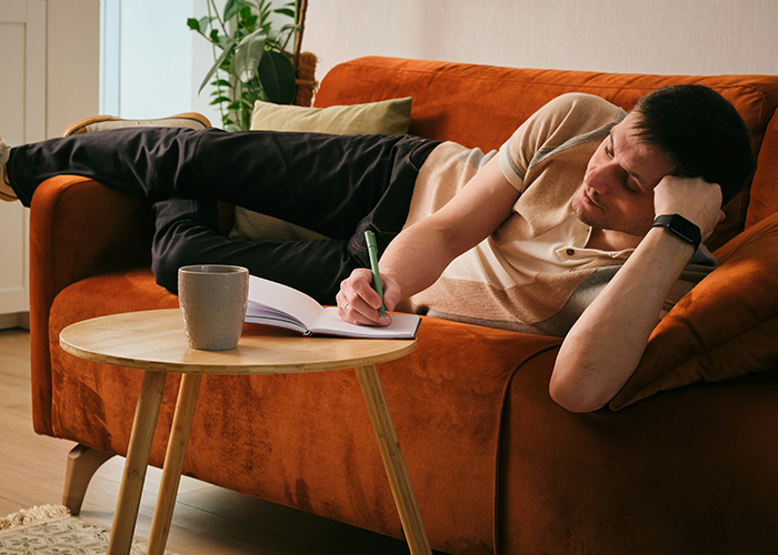 College student writing in notebook while lounging on orange couch in tidy shared apartment with plant in background. College student writing in notebook while lounging on orange couch in tidy shared apartment with plant in background.