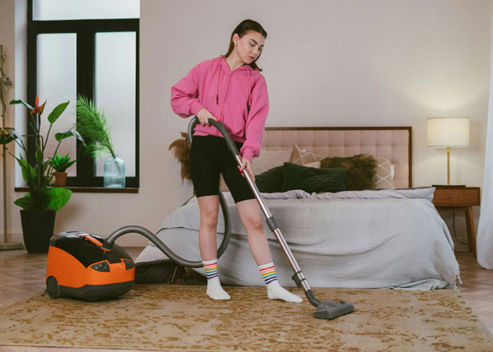 College student vacuuming bedroom carpet, wearing pink hoodie and striped socks, showing tidy roommate behavior. College student vacuuming bedroom carpet, wearing pink hoodie and striped socks, showing tidy roommate behavior.