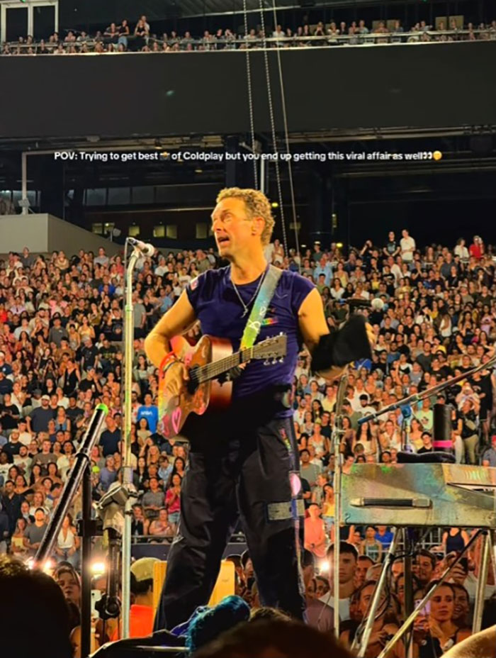 Chris Martin performing live on stage with guitar in front of a large crowd, capturing viral Coldplay couple behavior. Chris Martin performing live on stage with guitar in front of a large crowd, capturing viral Coldplay couple behavior.