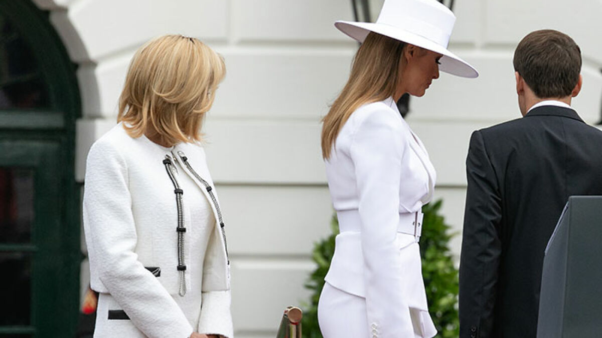 Melania Trumpu2019s curves highlighted in white outfit as she stands beside Brigitte Macron and a man outdoors near a building.