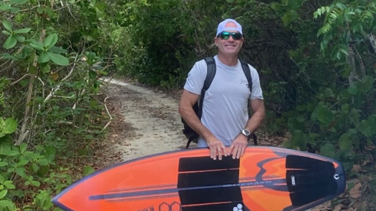 Florida surfer smiling, carrying an orange surfboard, surrounded by greenery after flying shark headbutt incident.