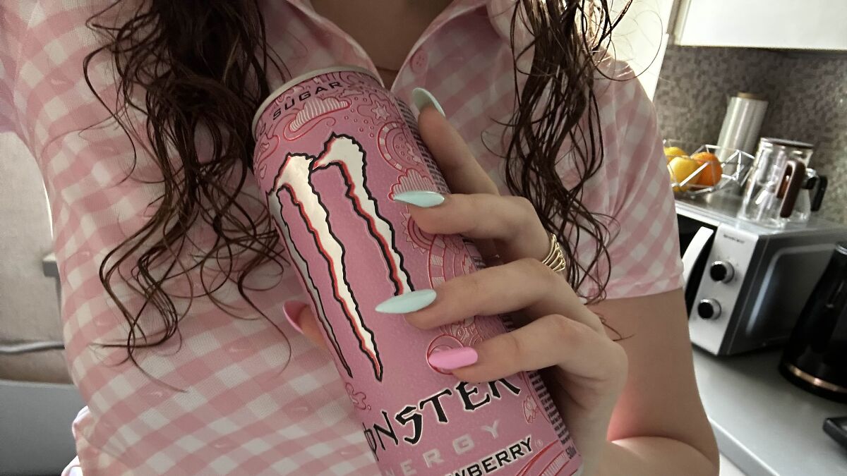 Person holding a pink Monster energy drink can with manicured nails in a kitchen setting.