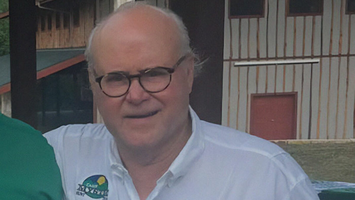 Director of girls camp and true hero, wearing glasses and a white shirt, in an outdoor setting near rustic buildings.