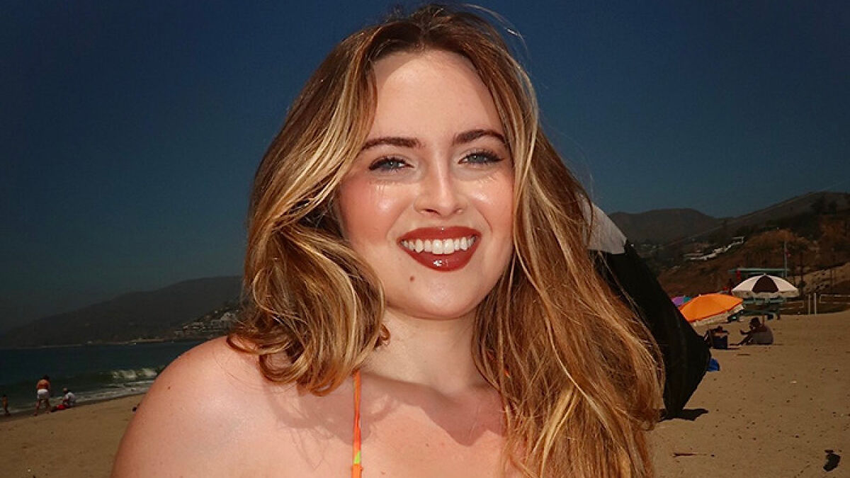 Plus-size model with long hair smiling confidently at the beach, embracing body positivity against fatphobic critics.