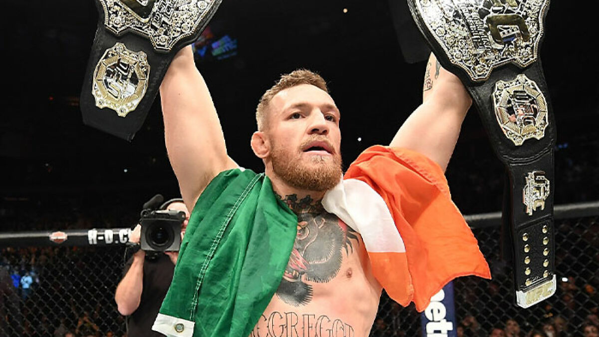 Conor McGregor holding UFC championship belts draped in Irish flag inside the octagon after a fight victory.