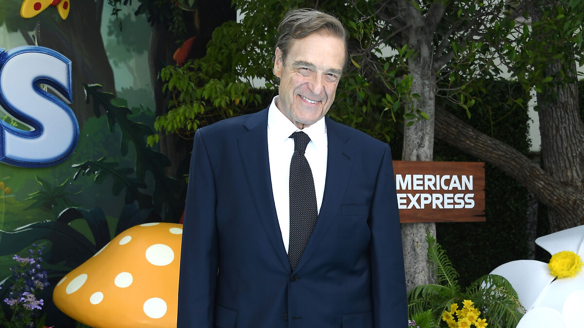 John Goodman smiling in a navy suit and tie at an outdoor event with greenery and colorful decorations behind him.