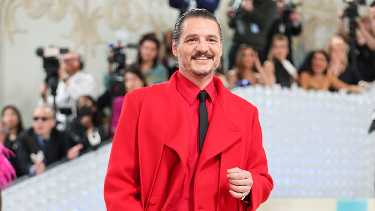 Pedro Pascal in a bright red suit smiling at an event with photographers and audience in the background.