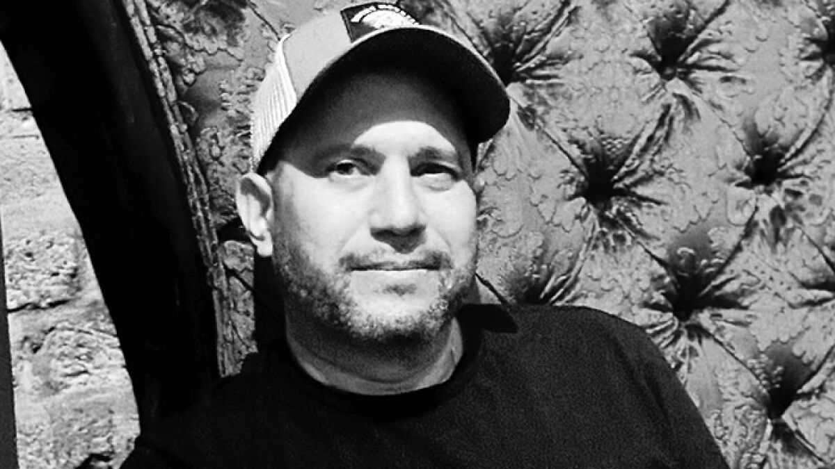 Paranormal investigator in casual wear and cap sitting against a patterned fabric backdrop during a tour.